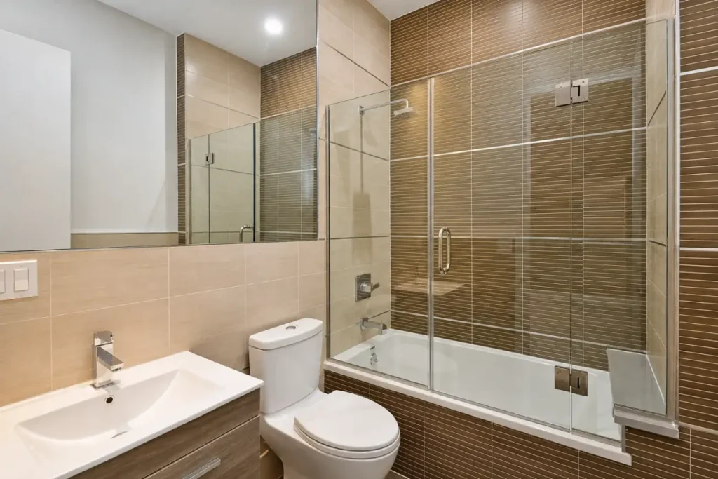 Completed modern bathroom with glass tub enclosure and warm tile in Hampton Bay