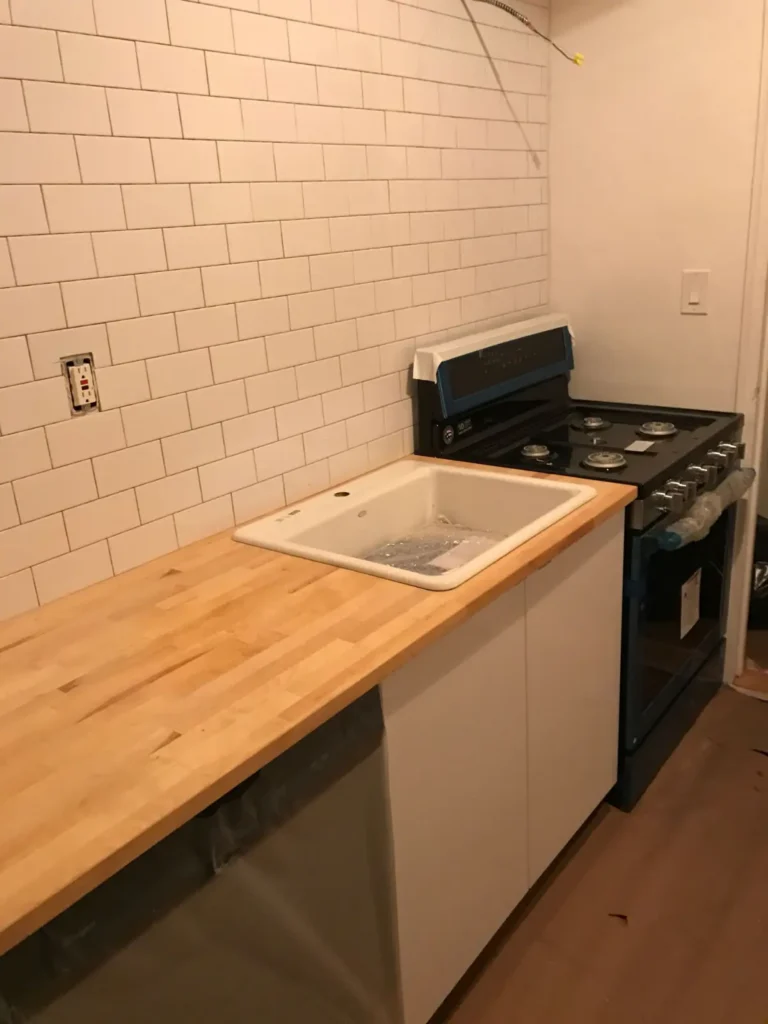 White galley kitchen cabinets, butcher-block counter, farmhouse sink, and subway tile during installation