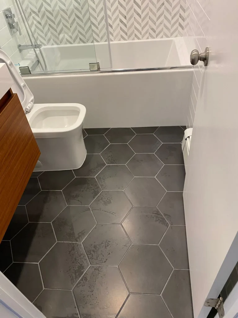 Dark charcoal hexagonal floor tile with floating wood vanity, toilet, tub, and chevron shower wall in a Downtown Brooklyn bathroom