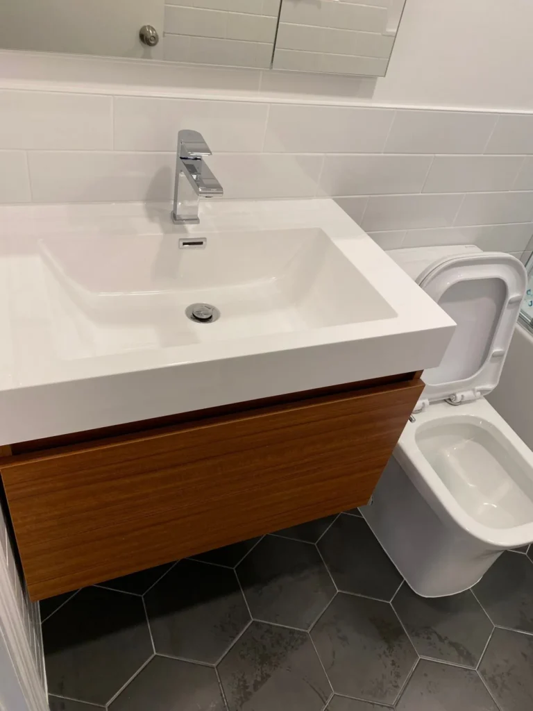 Wall-hung wood vanity with white integrated sink, mirrored cabinet, toilet, and dark hex floor tile in a compact Brooklyn bathroom