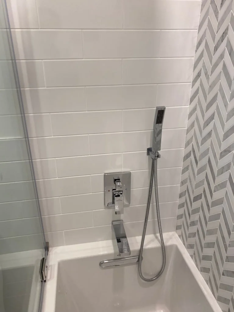 Chrome tub filler, shower control and handheld spray beside white subway tile and marble-look chevron tile in Downtown Brooklyn