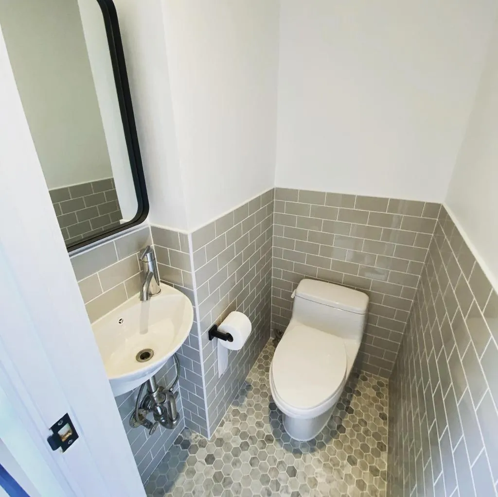 Compact Williamsburg powder room with wall-mounted corner sink, one-piece toilet, gray tile wainscot, and hex floor tile